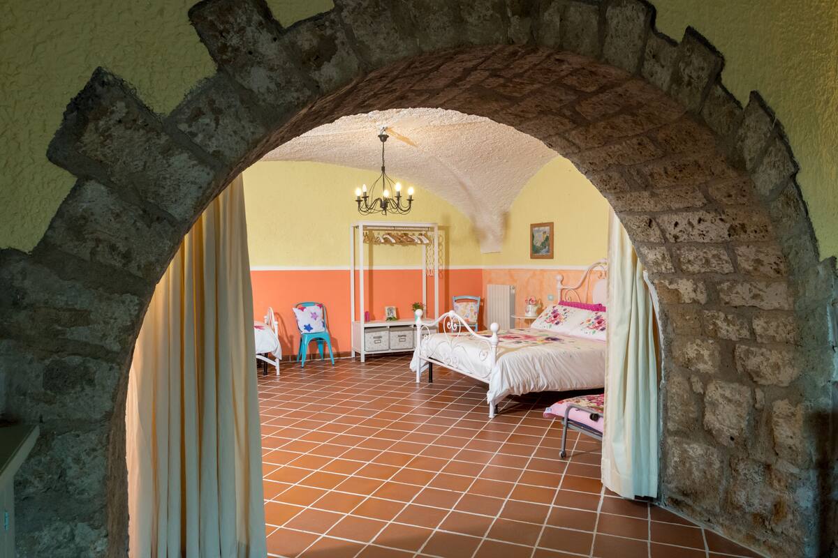 Um quarto com arco de pedra, uma cama, uma cadeira, cômoda e um lustre.