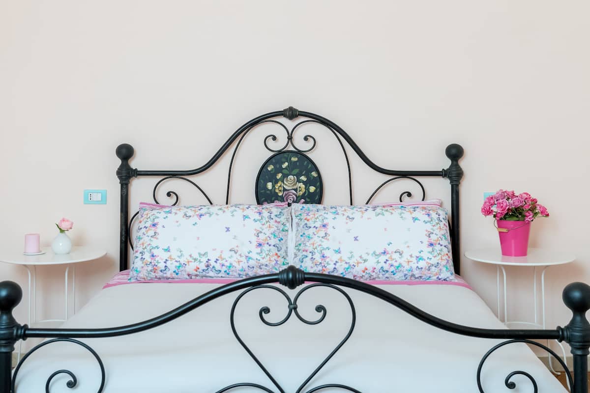 Uma cama com estrutura metálica preta, roupa de cama floral e almofada decorativa. Há duas mesas de cabeceira, uma com um vaso rosa e uma vela, e a outra com um balde rosa de flores.
