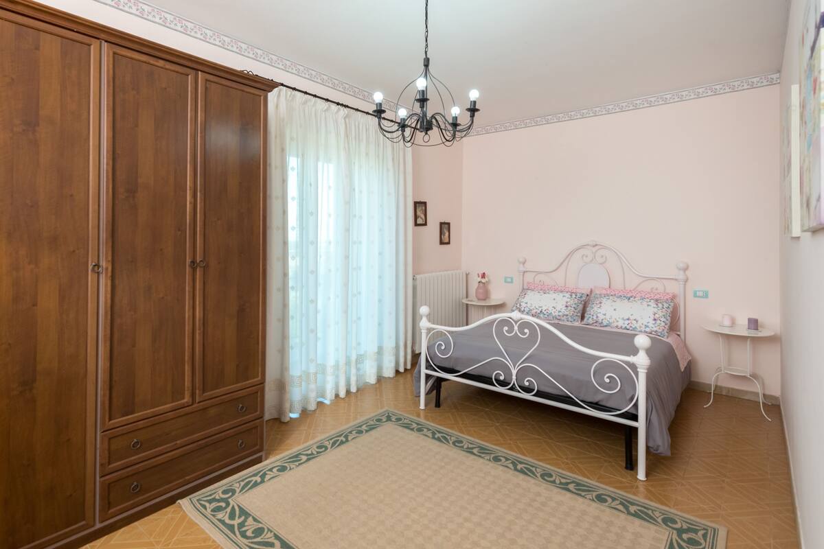 Quarto com um grande guarda-roupa de madeira, estrutura de cama de metal branco, um candelabro e uma pequena mesa de cabeceira.