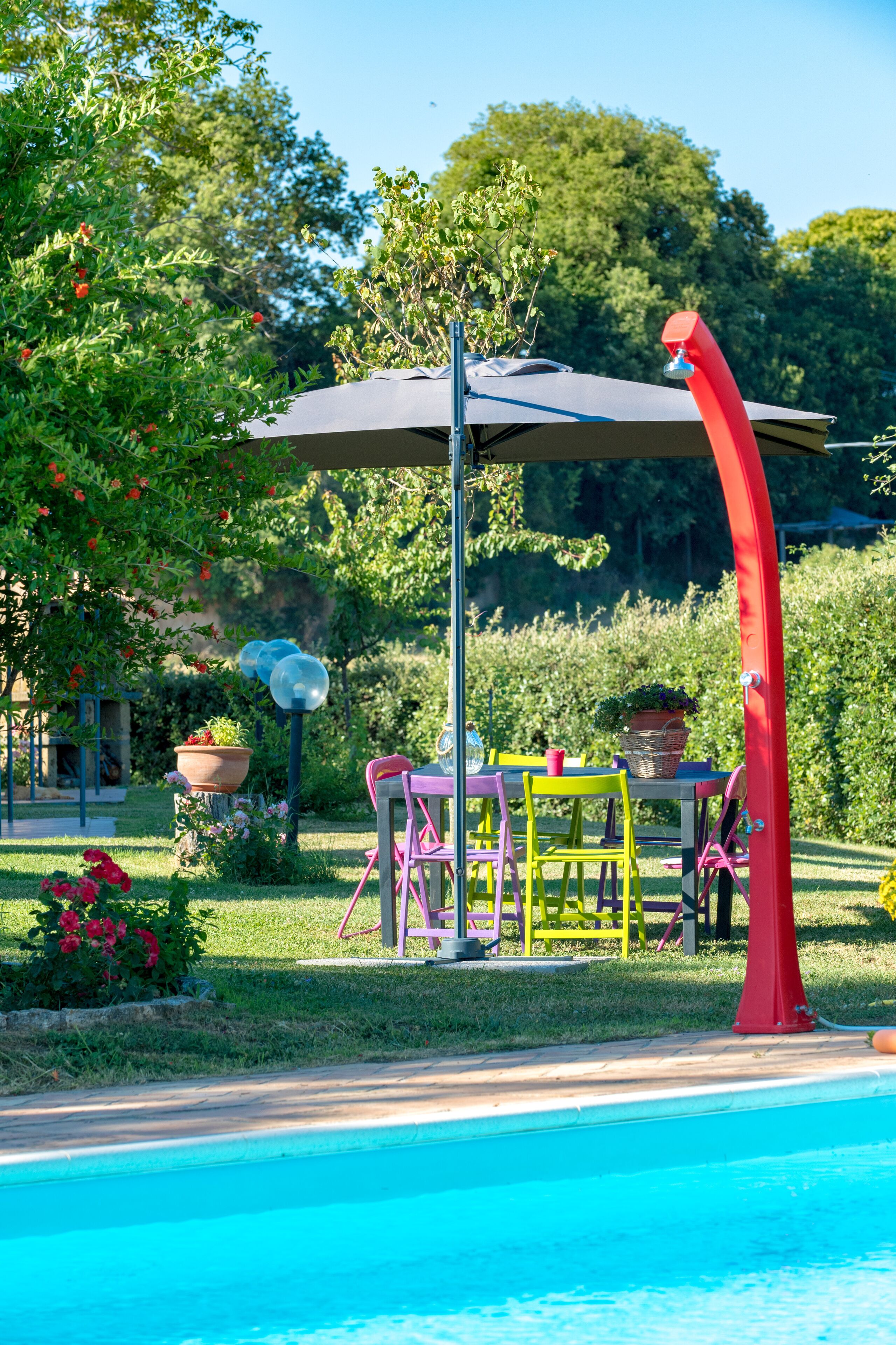 A poolside area with a table and chairs, surrounded by greenery and a clear sky.