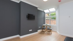 A modern room with a dark grey wall, a flat-screen TV mounted on the wall, a wooden dining table with chairs, and a large window providing natural light. - Crewe Rooms Ruskin Road (Crewe)