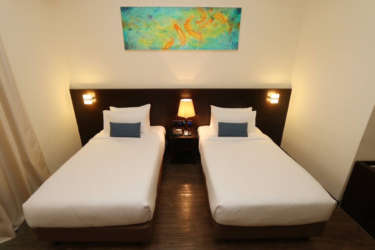A hotel room with two single beds, a nightstand with a lamp, and a painting on the wall.