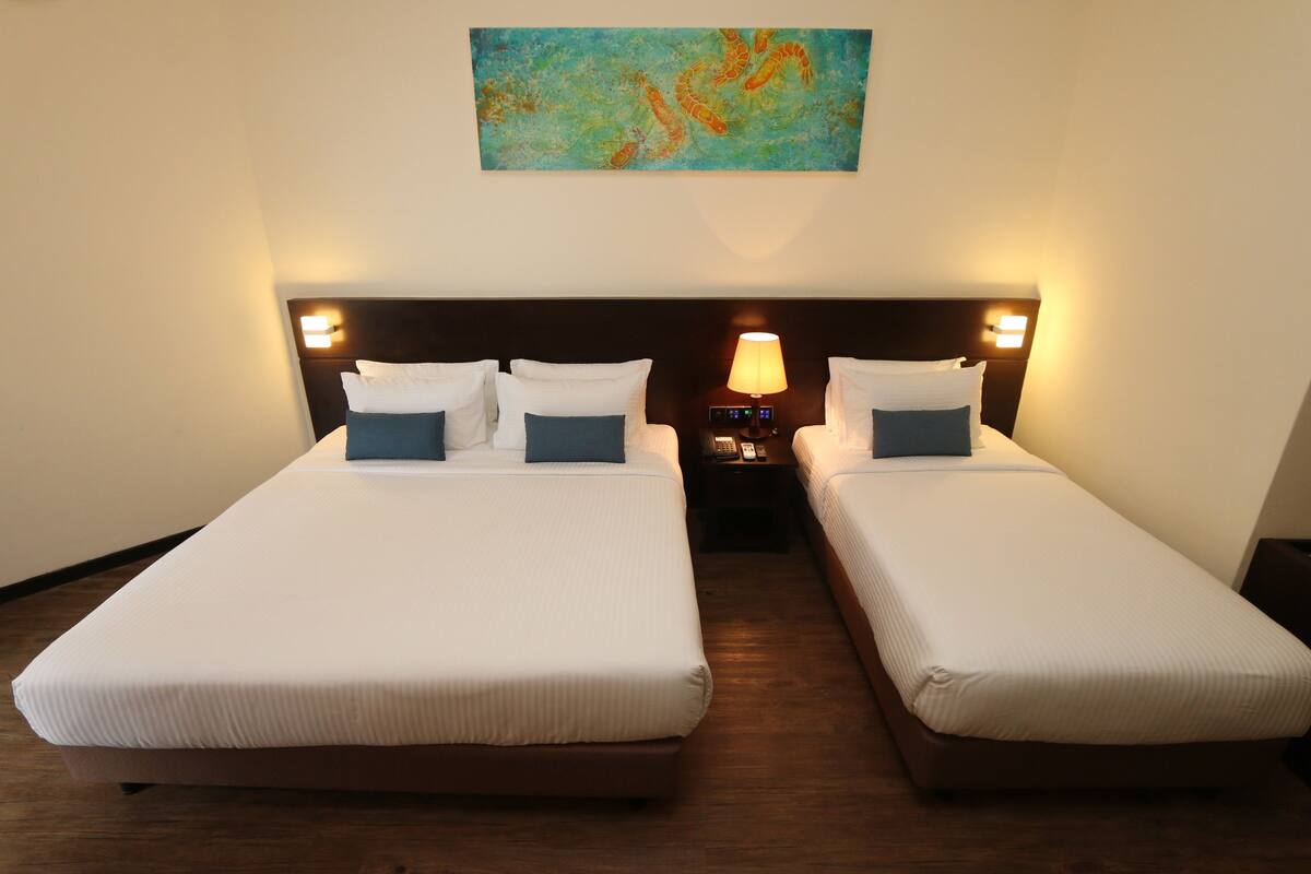A hotel room with two single beds, each with white linens and blue pillows, a nightstand with a lamp, and a painting on the wall.