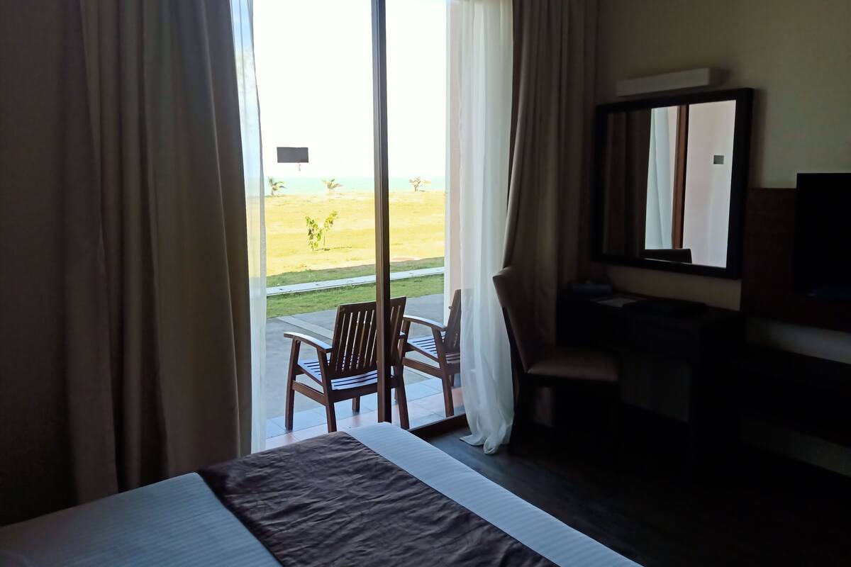 A hotel room with a bed, a desk, a chair, and a view of a beach through the window.
