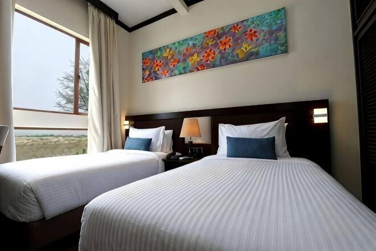 A hotel room with two beds, a nightstand with a lamp, a painting on the wall, and a window with curtains.