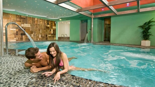 Ein Innenpool mit steinverkleideter Kante, einer Holzwand mit Mosaikmuster und einer grünen und orangefarbenen Decke.