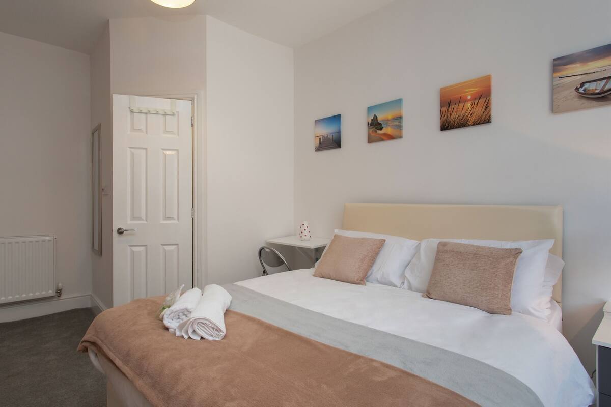 A neatly made bed with a beige headboard, a brown throw blanket, and a set of white towels. There are three framed pictures on the wall above the bed.
