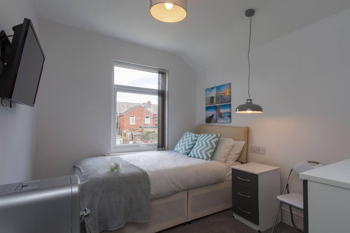 A bedroom with a bed, bedside table, desk, and a window with a view of a house.