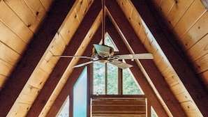 A wooden attic room with a sloped ceiling, a ceiling fan, and large windows offering a view of trees.