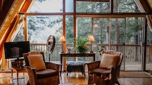 A cozy room with a large window, a television, wicker furniture, and a table with lamps and plants.