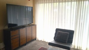 Una sala de estar con televisor de pantalla plana sobre un mueble de madera, sofá de cuero negro y cortinas de gasa.