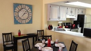 Un comedor con una mesa dispuesta para cuatro, sillas, un reloj en la pared y una cocina visible a través de un vano abierto.