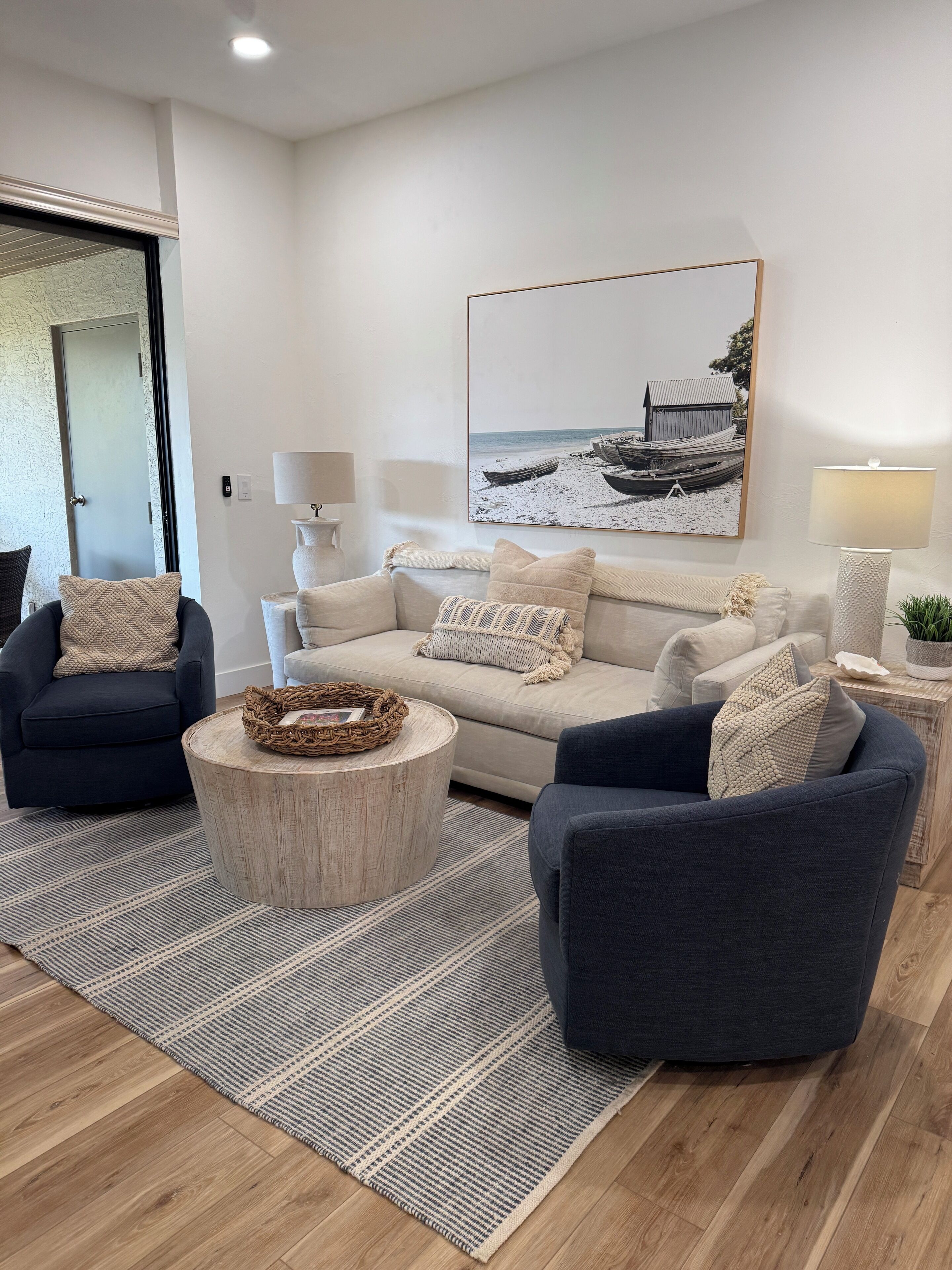 A modern living room with a beige sofa, two dark armchairs, a round coffee table, and a large framed picture of boats on a beach.