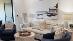 A modern living room with a beige sofa, two dark armchairs, a round coffee table, and a large framed picture of boats on a beach. - Luxury Beach Club- chairs, umbrellas, wifi, shelter included/ Pool, pickleball (Bonita Springs)