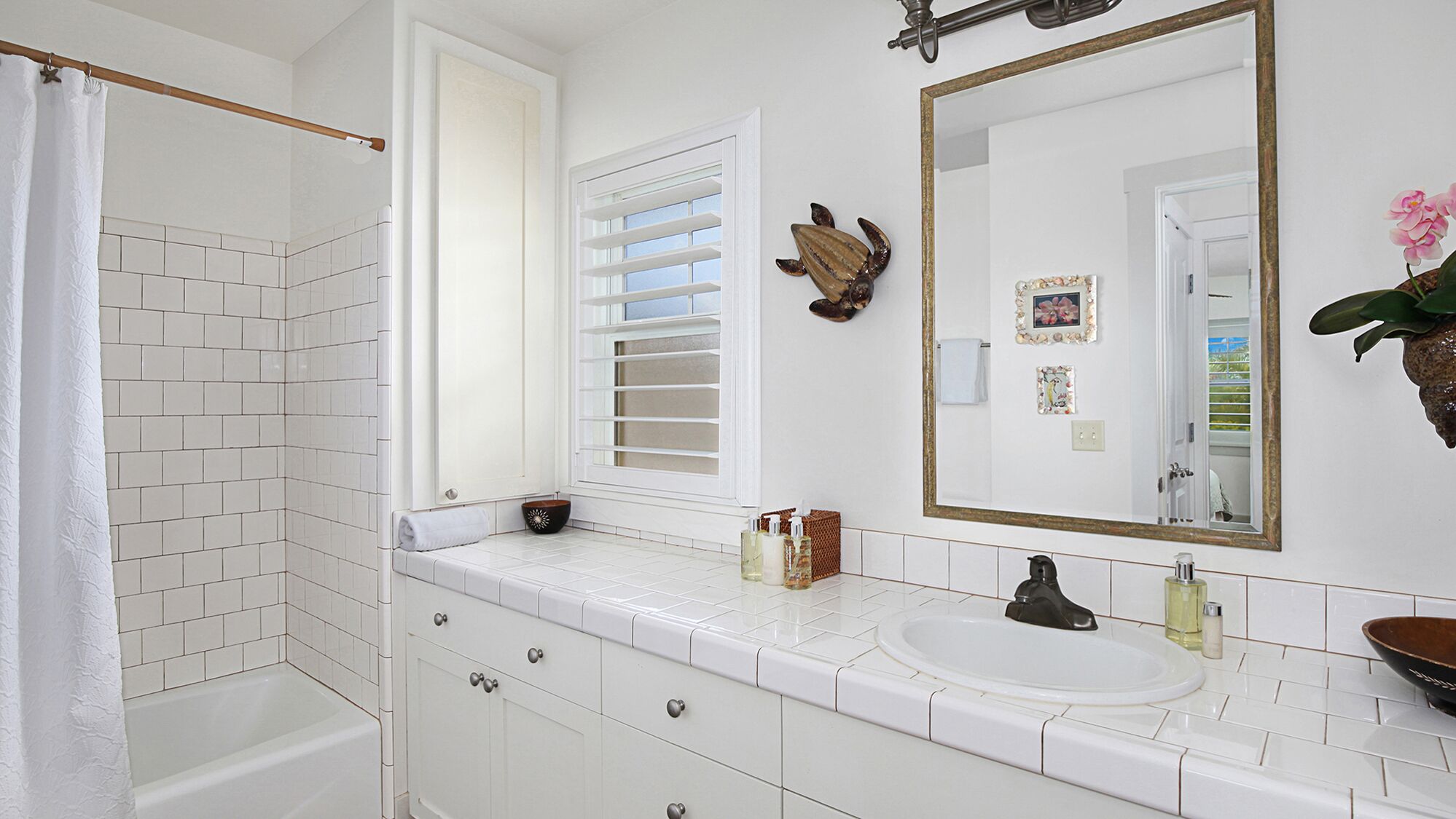 A bathroom with a white tiled shower, a window with blinds, a sink with toiletries, and a mirror with a decorative frame.