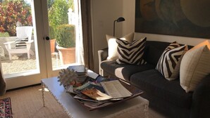 Un saló amb un sofà fosc, coixinets blancs, una taula de centre amb revistes i una vista a un jardí a través d'una porta corredissa.