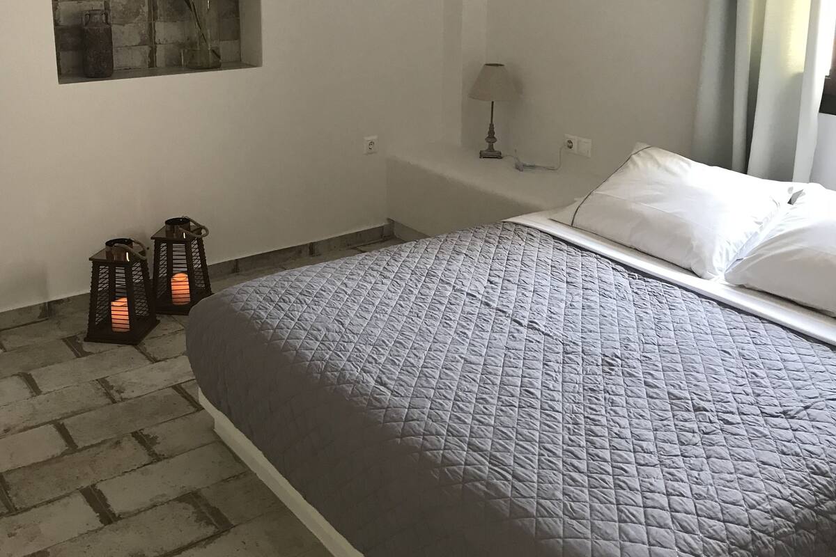 Quarto com cama, janela com vista do céu, ar-condicionado fixado na parede e pequena mesa com abajur.