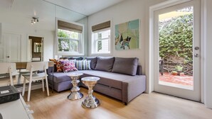 A modern living room with a grey sofa, white chairs, and a dining table. There is a large window with blinds, a door leading outside, and a mirror on the wall. - Beachcombers Hideaway, Dog Friendly, Enclosed Patio, 1 Block to Beach (La Jolla)