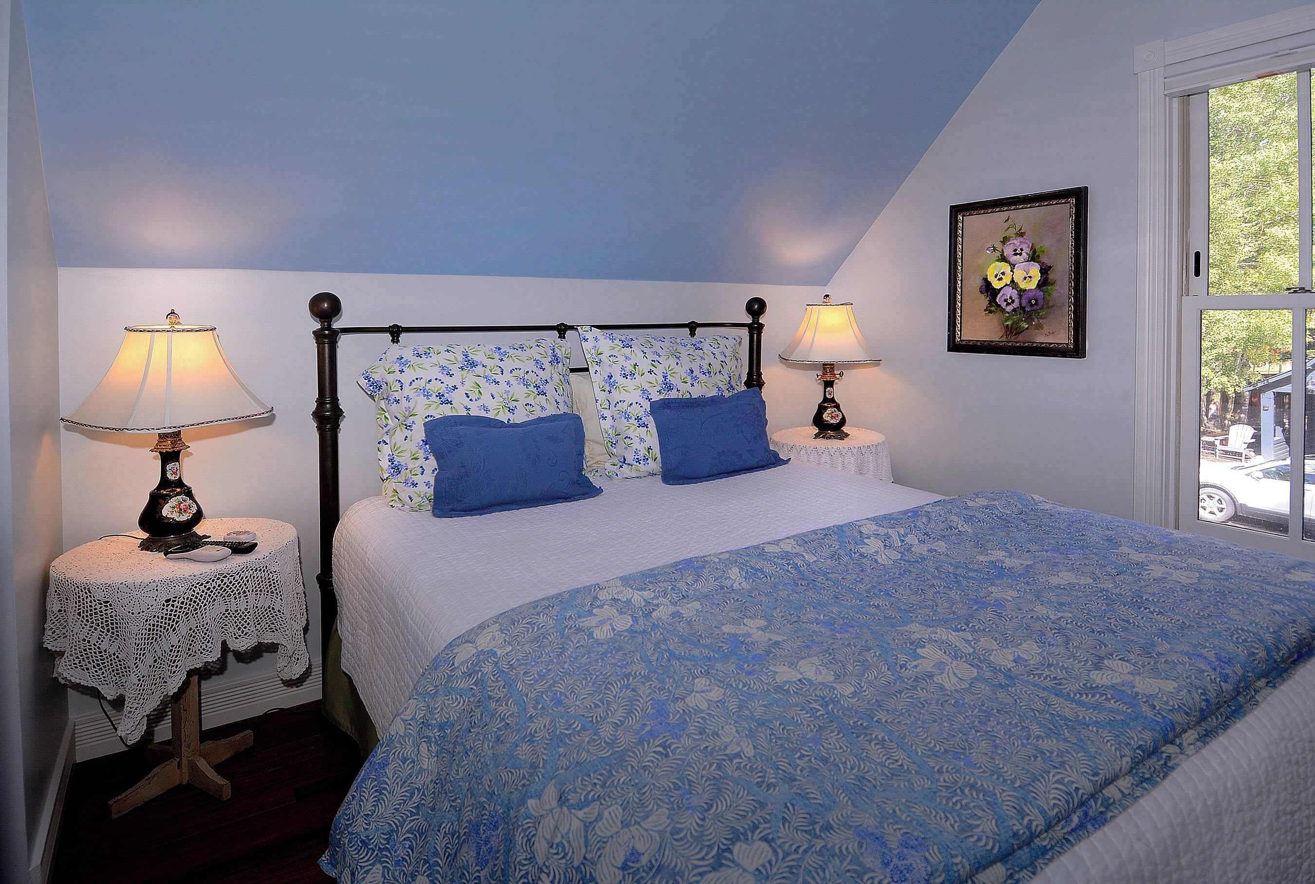 A bedroom with a bed, two bedside tables, two lamps, a floral wall decoration, and a window with a view.