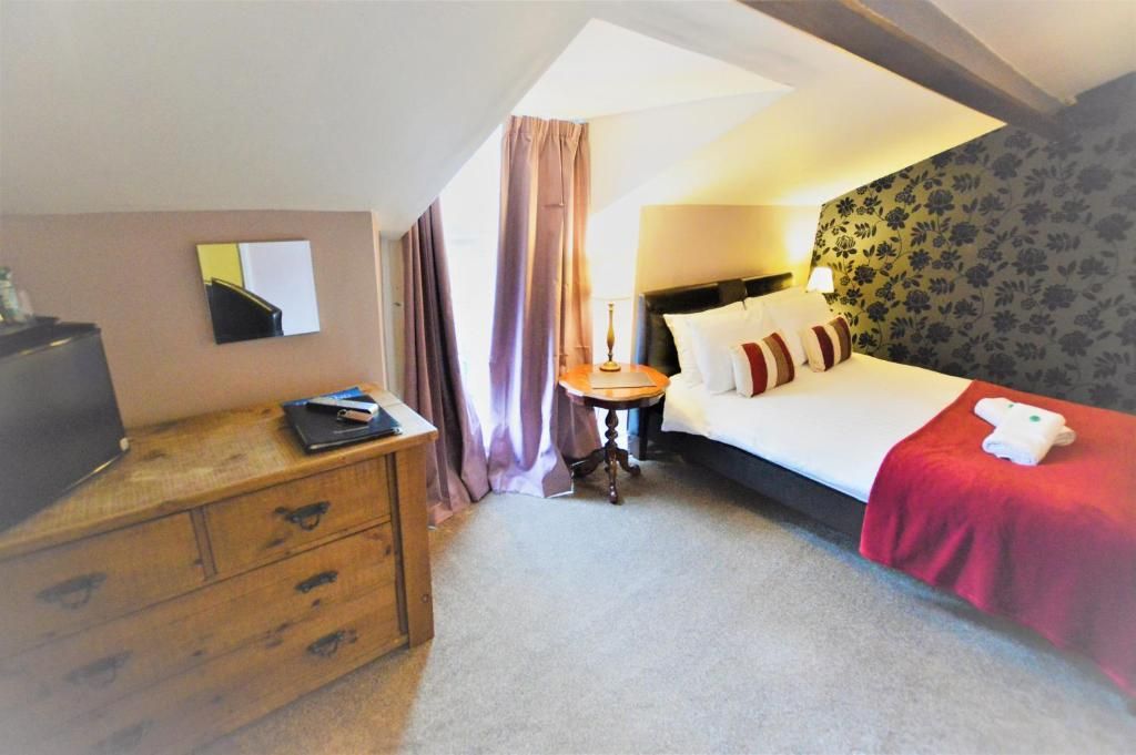 A hotel room with a bed, a wooden dresser, a small table with a lamp, and a television on the dresser.