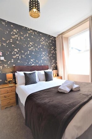 A hotel room with a bed, a nightstand, a lamp, and a window with curtains. - All Seasons Rest (Windermere)