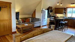 A cozy living room with a sofa, coffee table, and a wooden bench. A kitchen with a refrigerator, microwave, and bar stools is visible. - Stage Road Estate private studio apartment. (Columbia Falls)