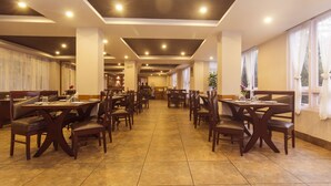 Un restaurant spacieux, doté de nombreuses tables et chaises, de hauts plafonds avec des luminaires encastrés et de grandes fenêtres ornées de rideaux légers.