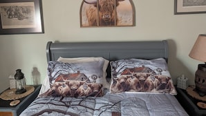 Una cama con dos almohadas decoradas con motivos de vaca de las Tierras Altas, un cuadro enmarcado de un toro y una lámpara en la mesita de noche.