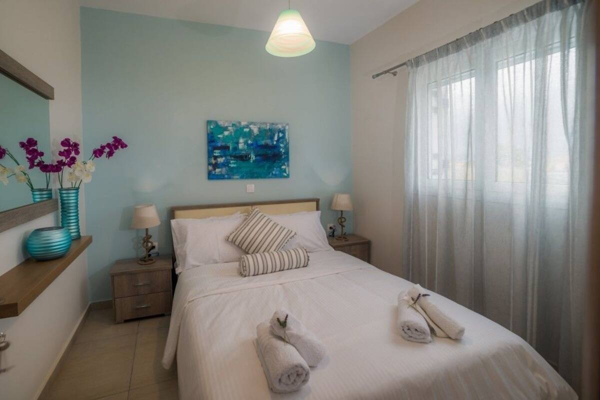 A hotel room with a bed, two bedside tables, a nightstand, a wall-mounted painting, and a window with sheer curtains.