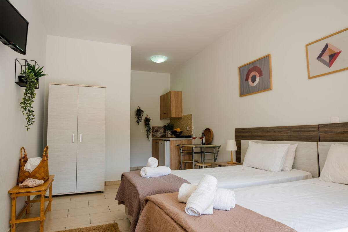 A hotel room with two beds, a wooden headboard, a small table with a plant, and a kitchenette in the background.