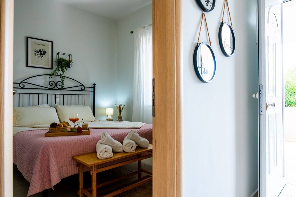 A bedroom with a bed, bedside table, and a bench with towels. There are wall decorations and a window with curtains.