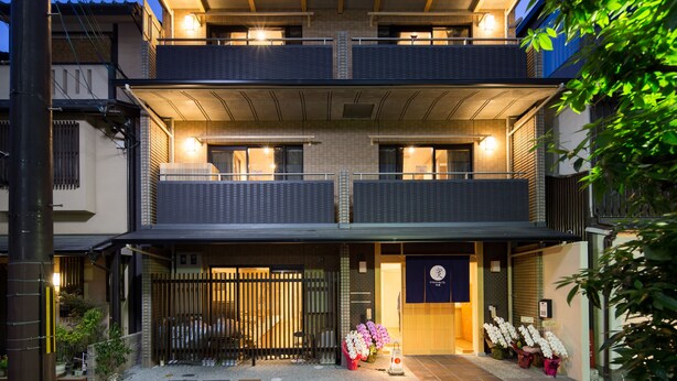 A two-story building with a balcony on the upper floor, a small entrance area with a sign, and potted plants outside.