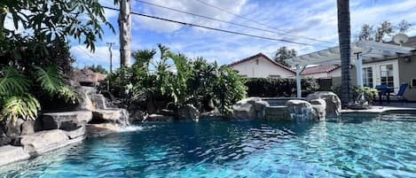 En klar och inbjudande pool med ett vattenfall som kaskaderar ner över stenar, omgiven av frodig grönska och ett hus i bakgrunden.