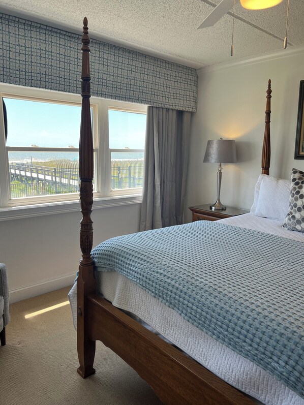 Un dormitorio con cama con dosel, vistas al mar a través de una ventana, una lámpara en una mesita de noche y un ventilador de techo.