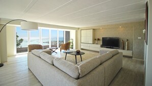 A modern living room with a large sofa, a television, and a view of the ocean through large windows.