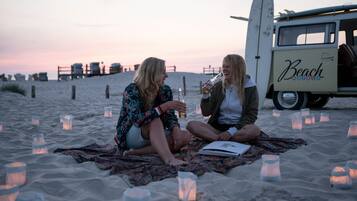 Två personer sitter på en filt på stranden, med en surfbräda och ett fordon i bakgrunden som har texten ”Beach” på sig.