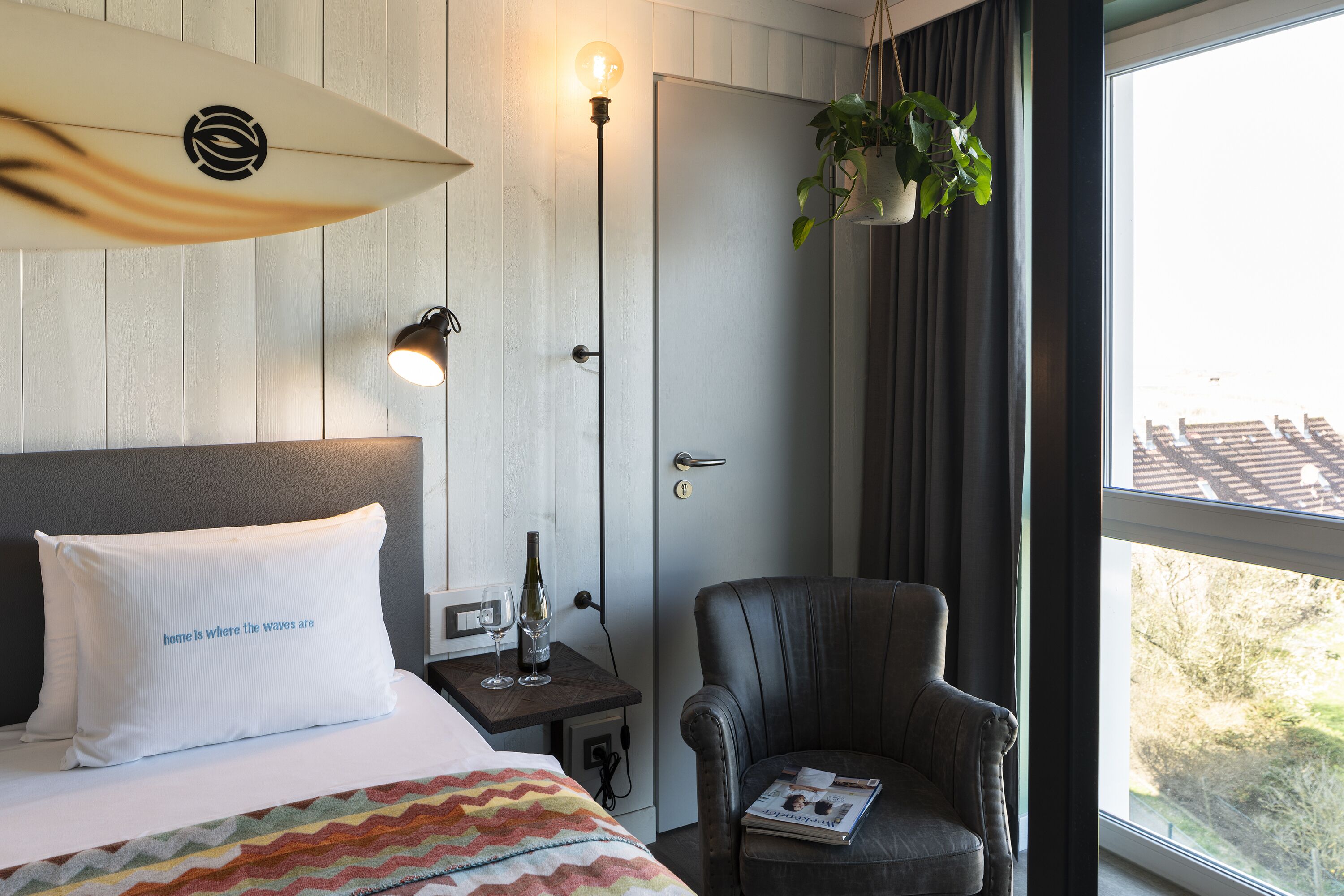 A hotel room with a bed, a nightstand, a reading chair, a plant, and a window with a view.