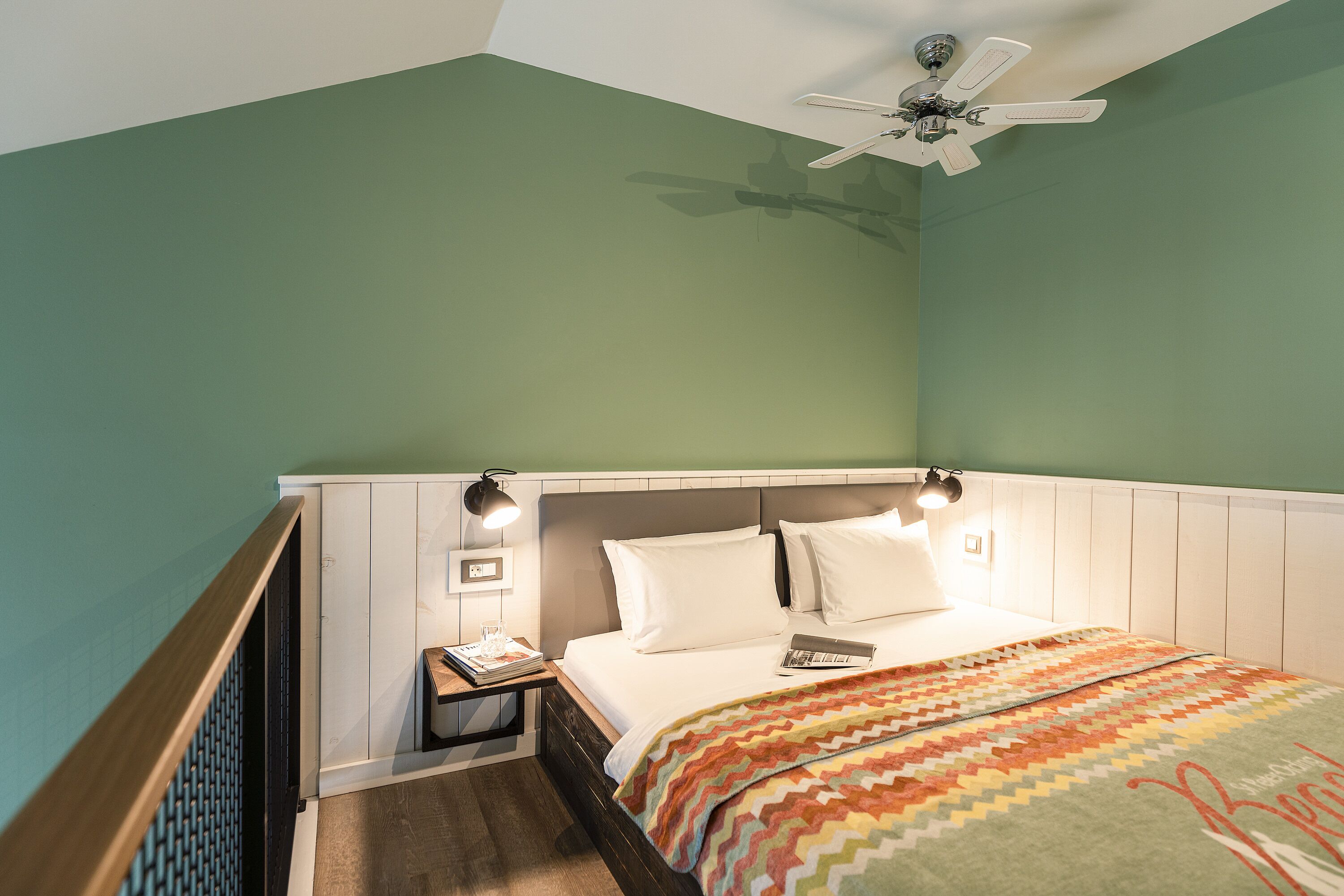 A hotel room with a bed, a nightstand, a ceiling fan, and a wall-mounted lamp.