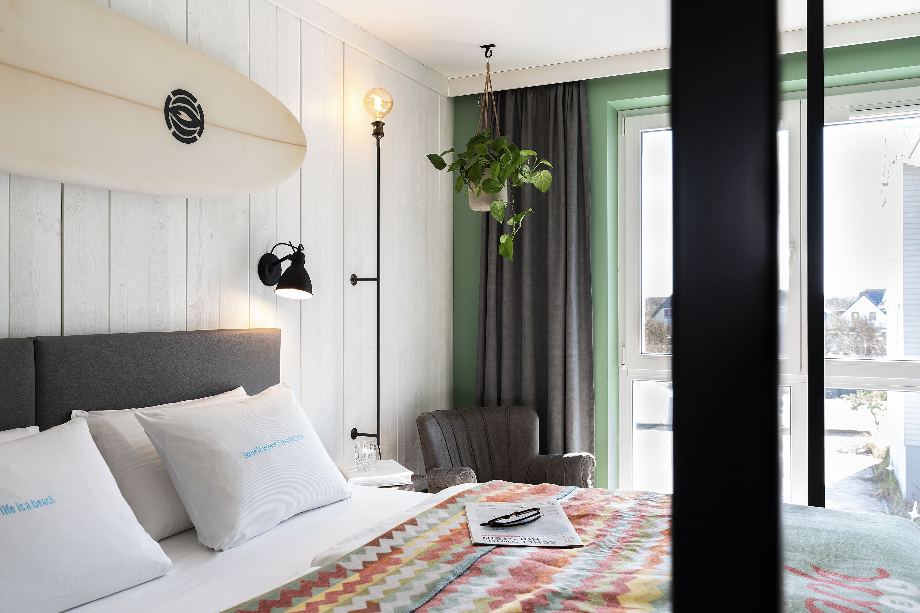 Ein Hotelzimmer mit Bett, Surfbrett-Dekoration, hängender Pflanze, Stuhl und Fenster mit Blick.