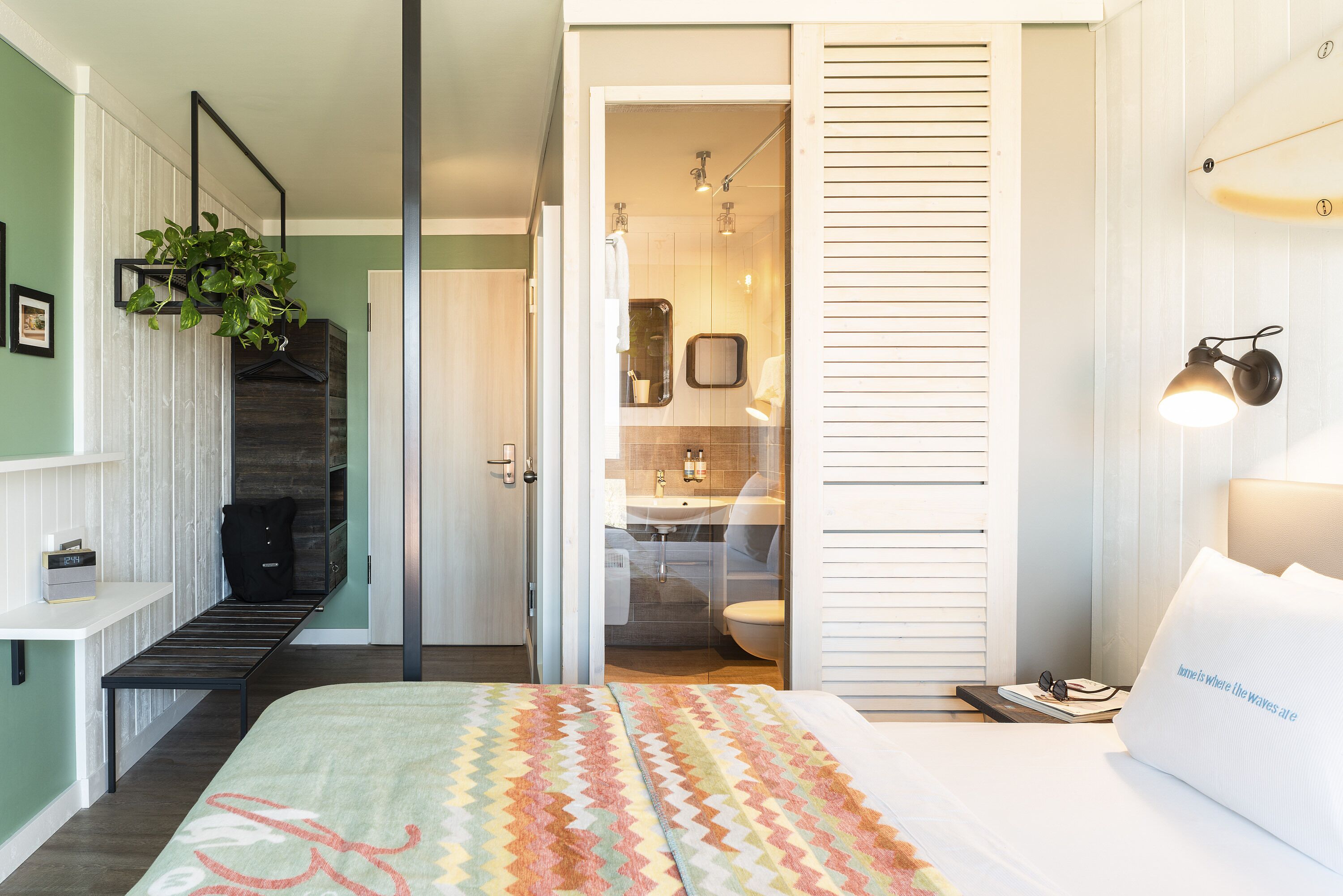 A bedroom with a bed, a bedside table, a hanging plant, a bathroom with a sink and toilet, and a sliding door leading to a shower area.