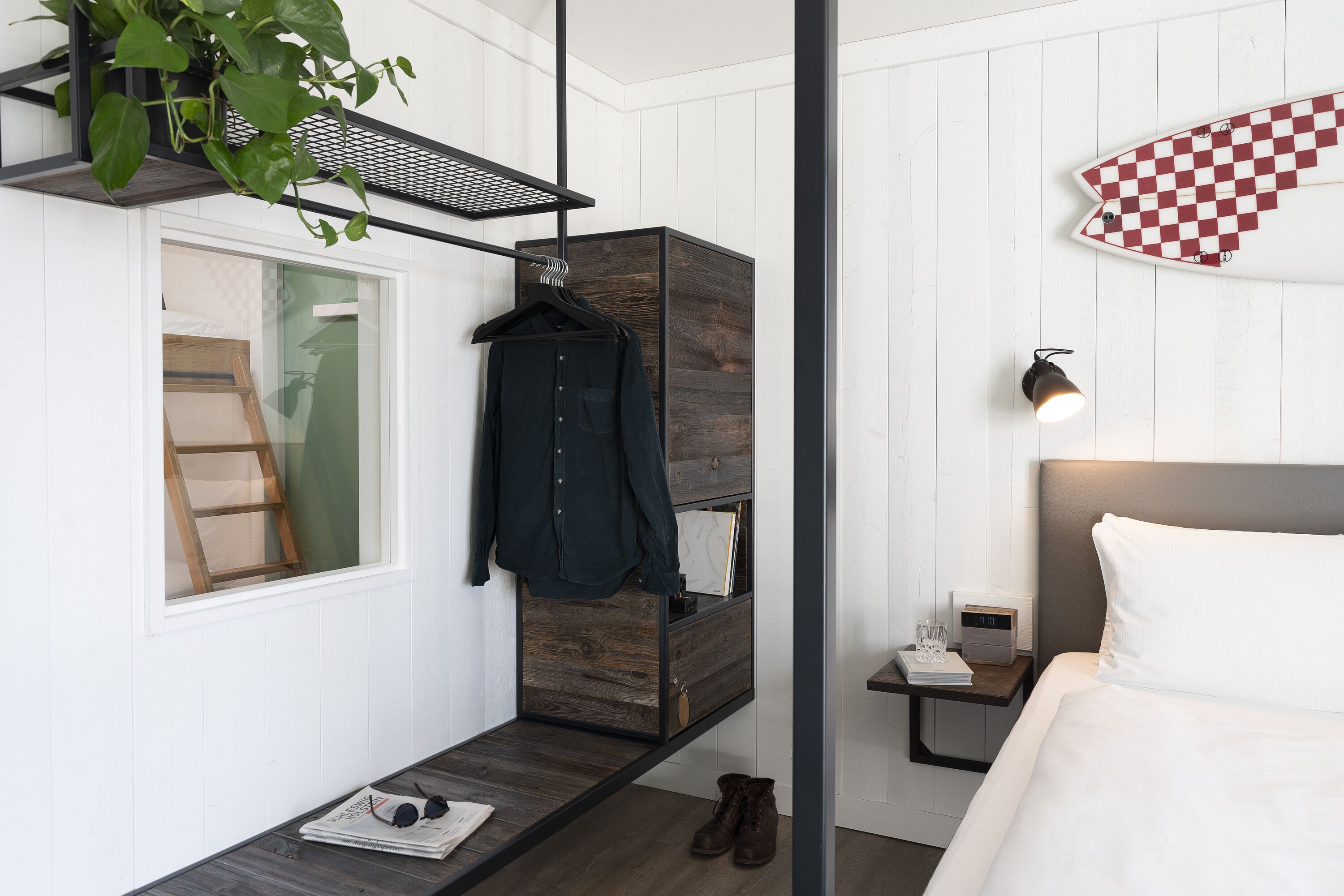 A modern bedroom with a wooden dresser, a hanging coat rack, a bedside table with a lamp, and a checkered wall decoration.