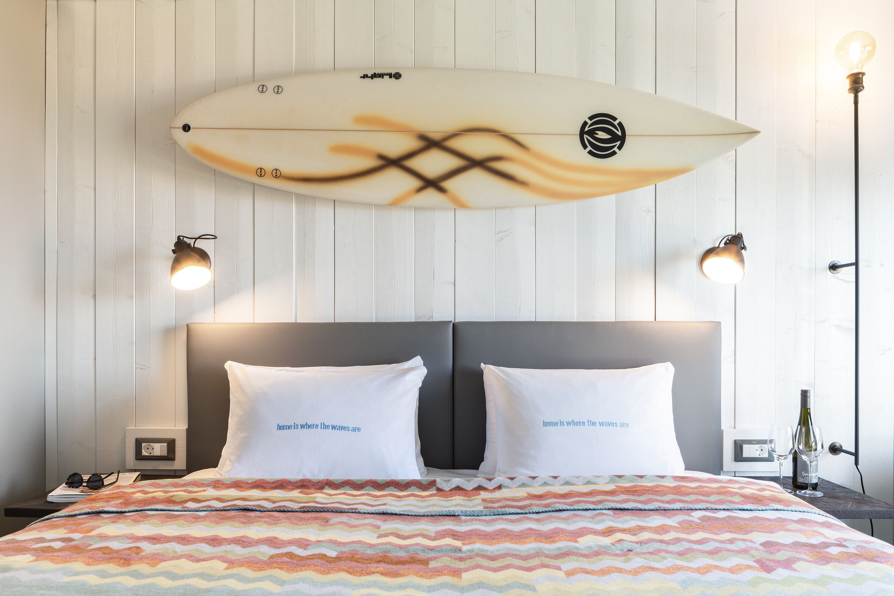Una cama con una colcha de colores con estampado en zigzag, dos almohadas blancas con texto, una decoración de tabla de surf sobre la cama y dos lámparas fijadas a la pared.