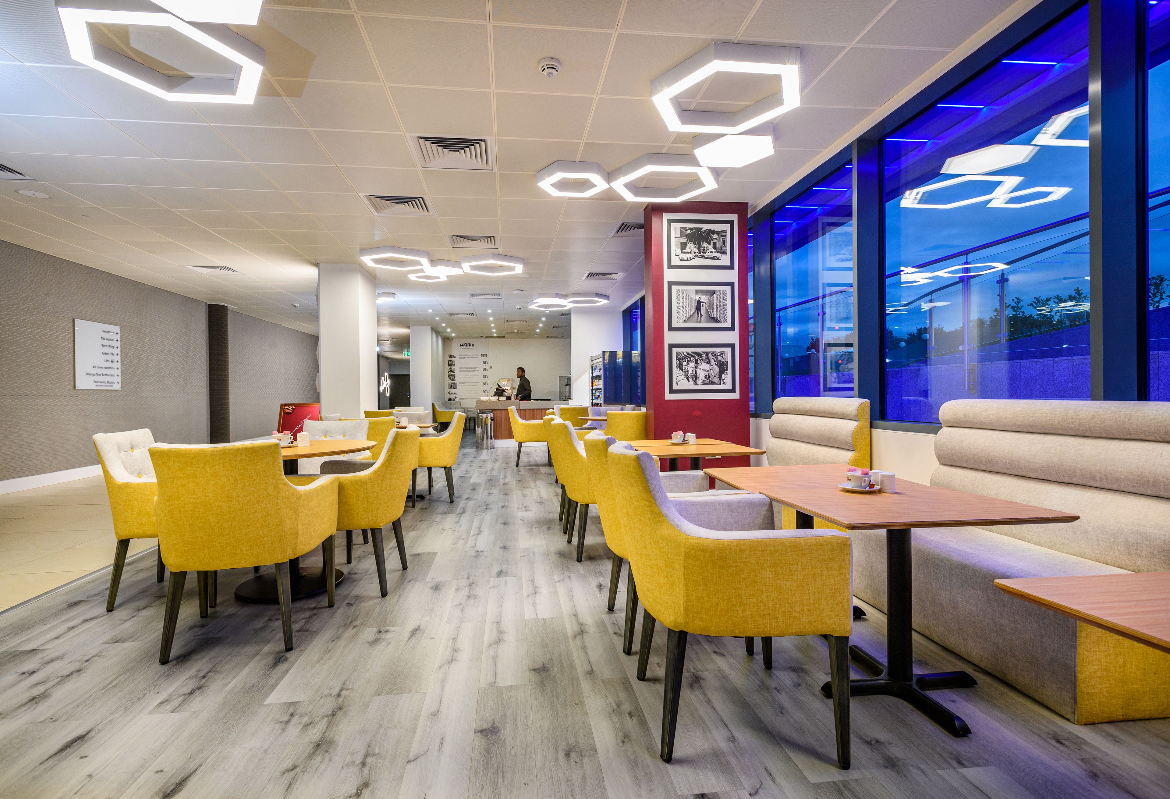 A modern restaurant with yellow chairs and tables, a blue-lit wall, and a seating area with a sofa and benches.