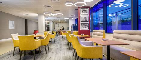 Un restaurant moderne avec des chaises et des tables jaunes, un mur éclairé en bleu et un espace détente comprenant un canapé et des bancs.