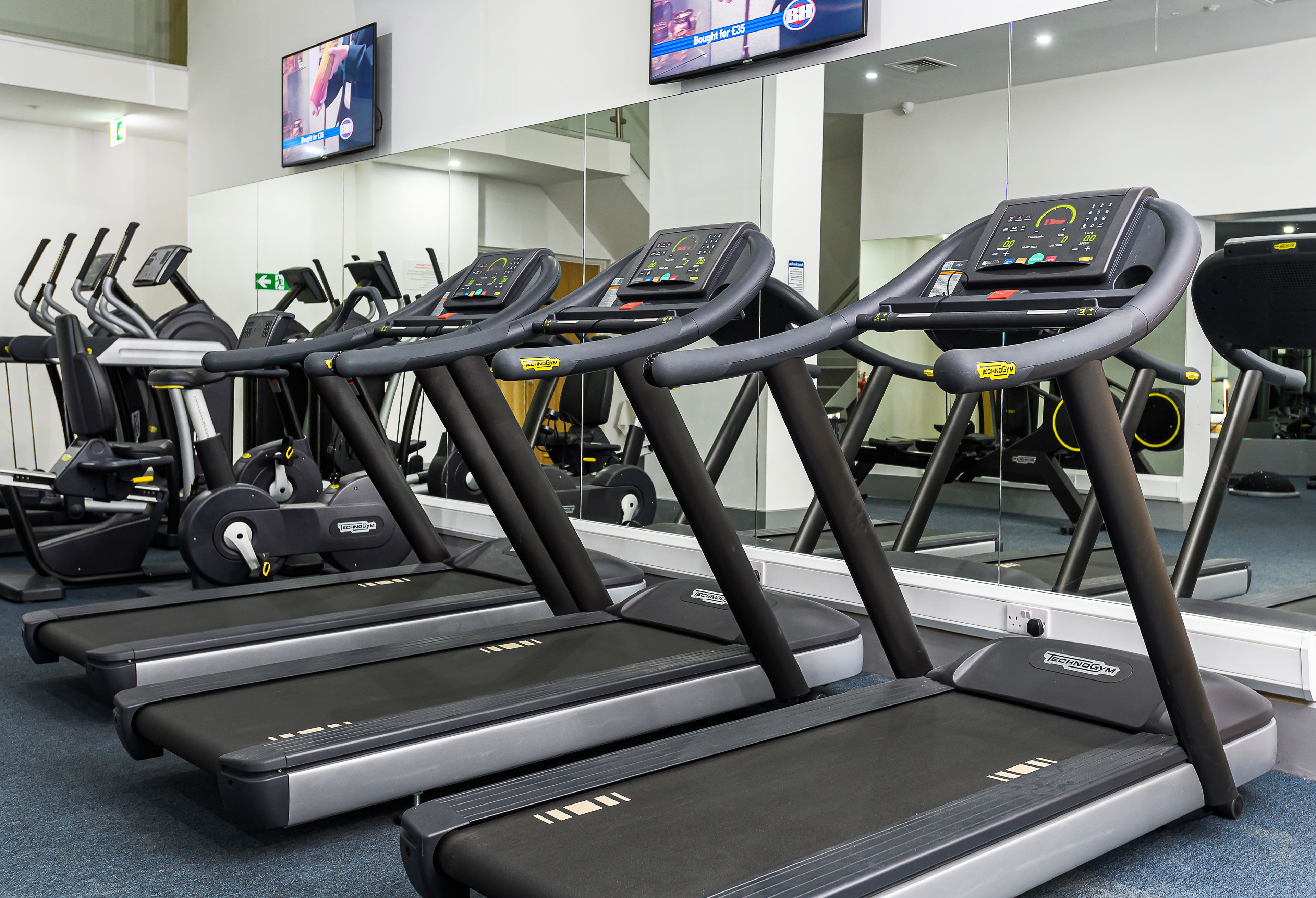 A row of treadmills in a gym with a TV mounted on the wall.