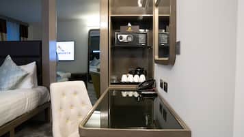 A hotel room with a bed, a seating area with a cushioned chair, a minibar, a coffee maker, and a telephone.