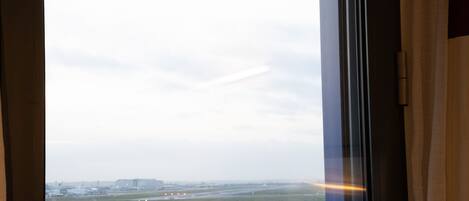 A view through a window of a parking lot with multiple rows of parked cars, a distant airport, and a cloudy sky.