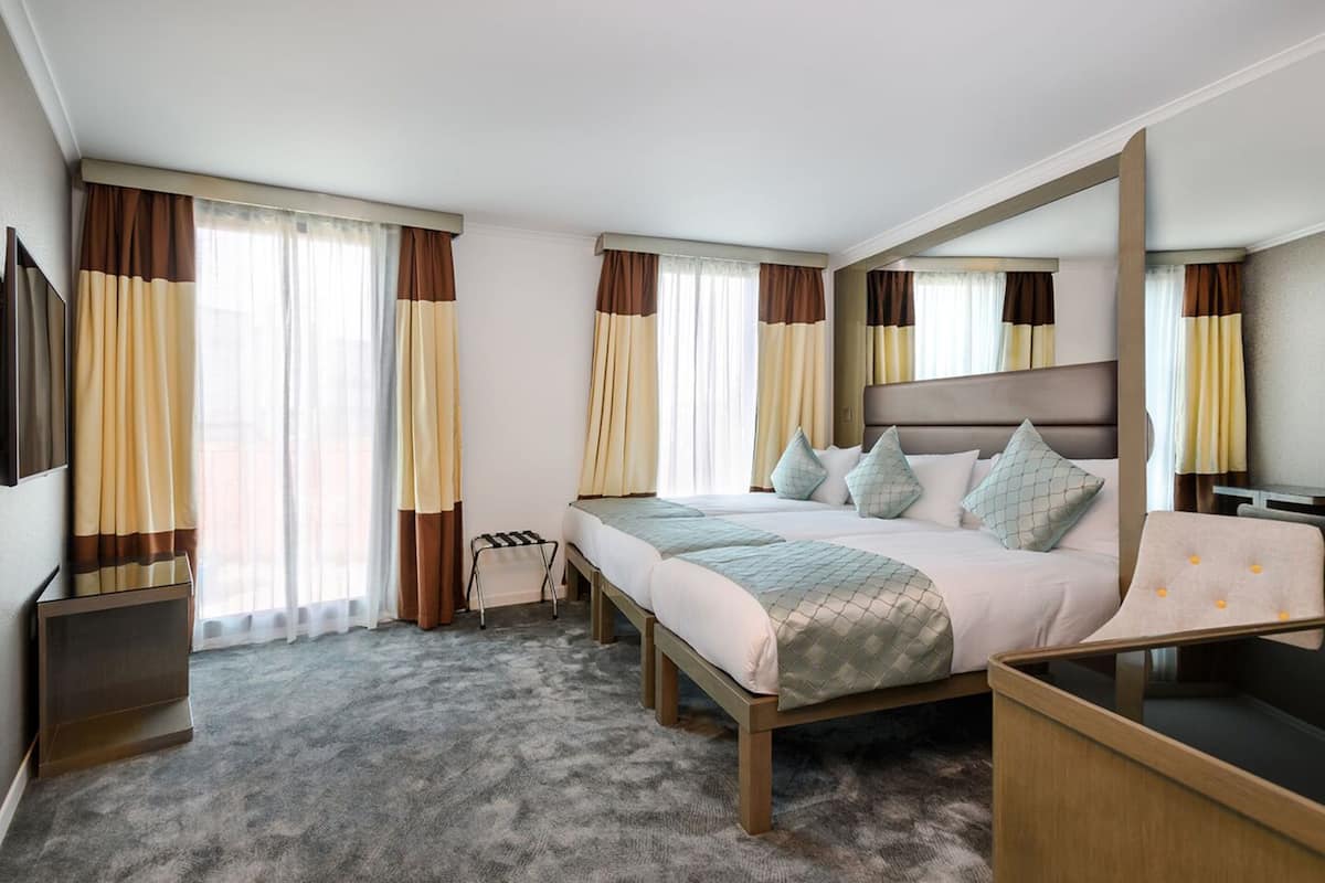 A hotel room with a large bed, a desk, and a chair. There are curtains on the windows, and a flat-screen TV on the wall.