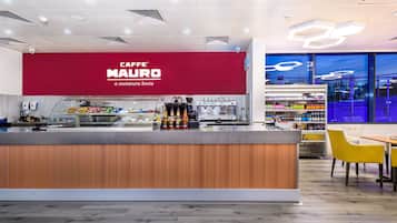 Un café moderno con un llamativo letrero rojo, una barra con bebidas y una zona de estar con sillas amarillas.