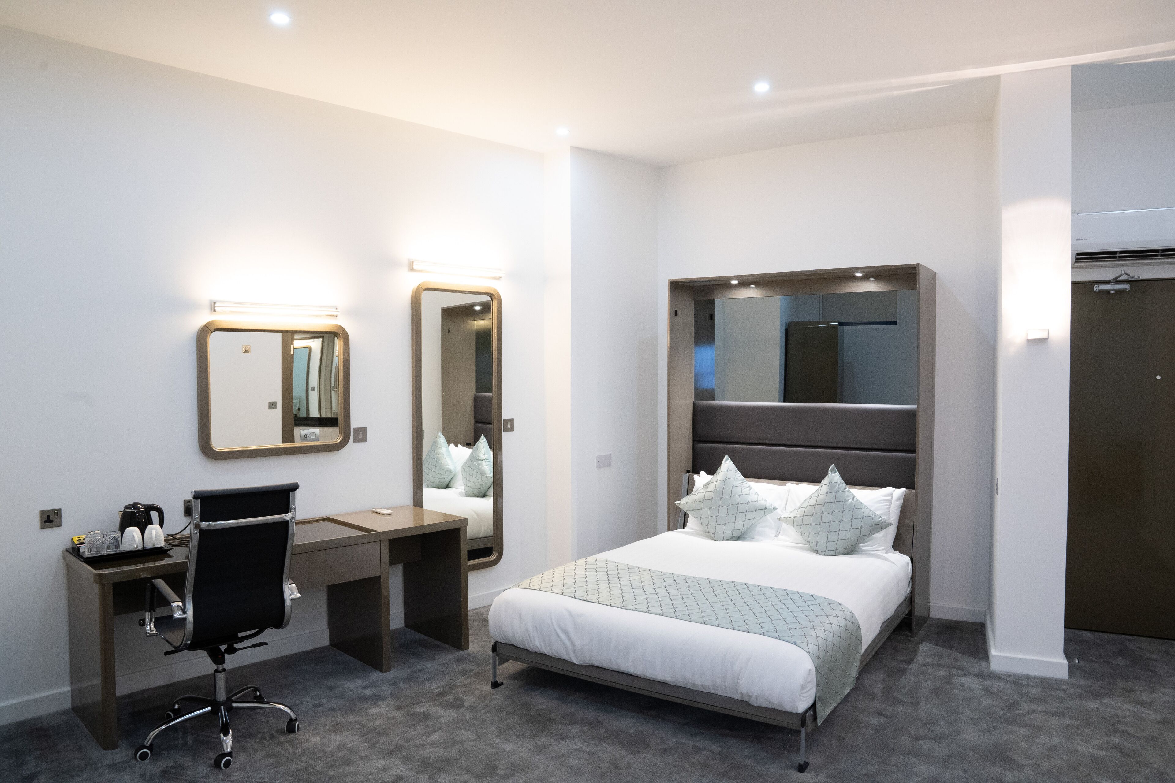 Una habitación de hotel moderna con una cama, un escritorio con silla, un espejo grande y un armario.